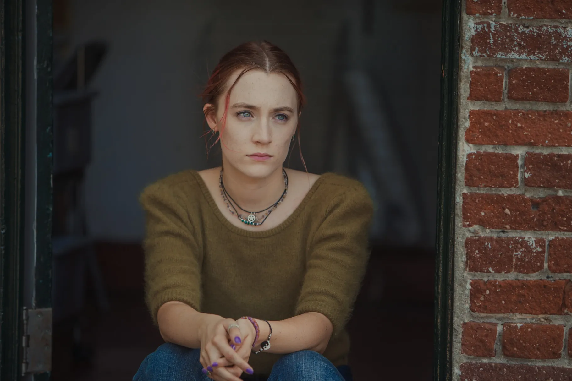 Lady Bird stares into the distance, wearing a green sweater and jeans.