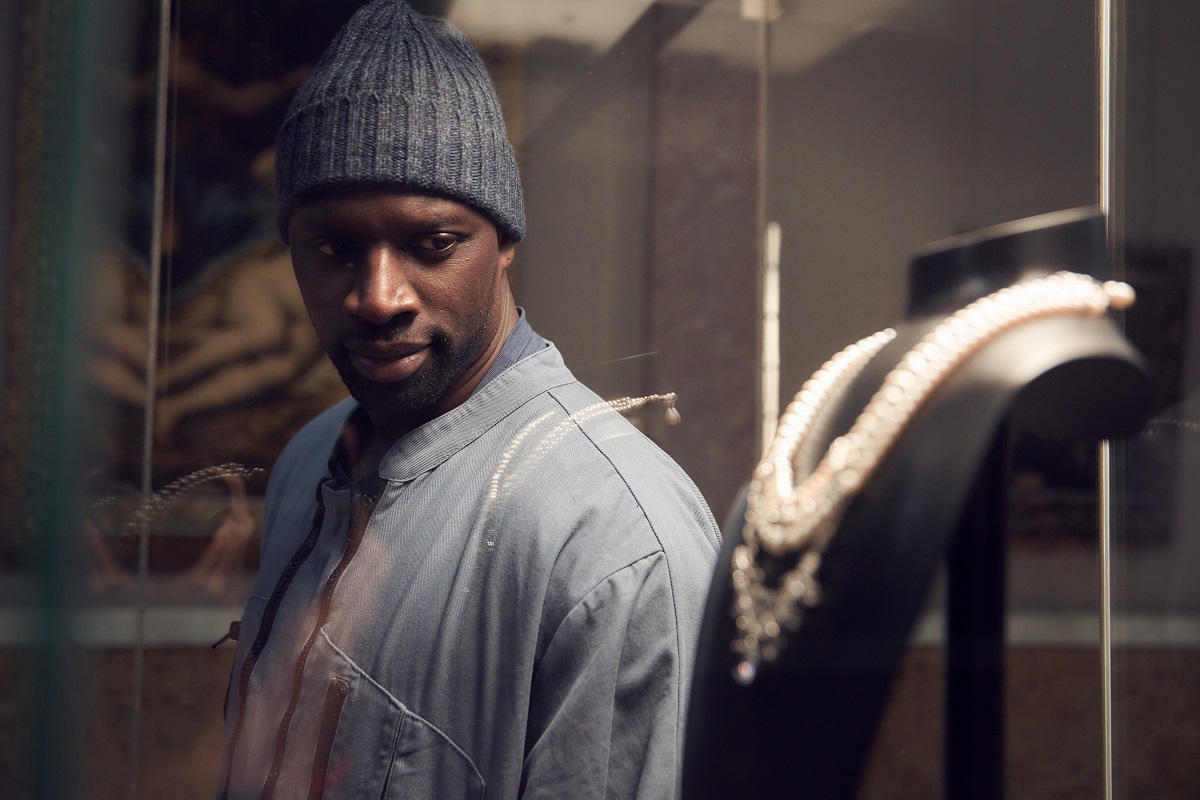 Assane played by Omar Sy is staring into a glass cabinet with jewels in. 