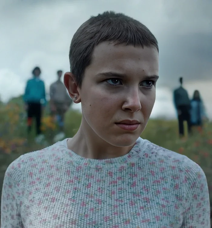 Eleven from Stranger Things Wiki. She has short hair and looks angry. There are people out of focus behind her on what appears to be a hill.