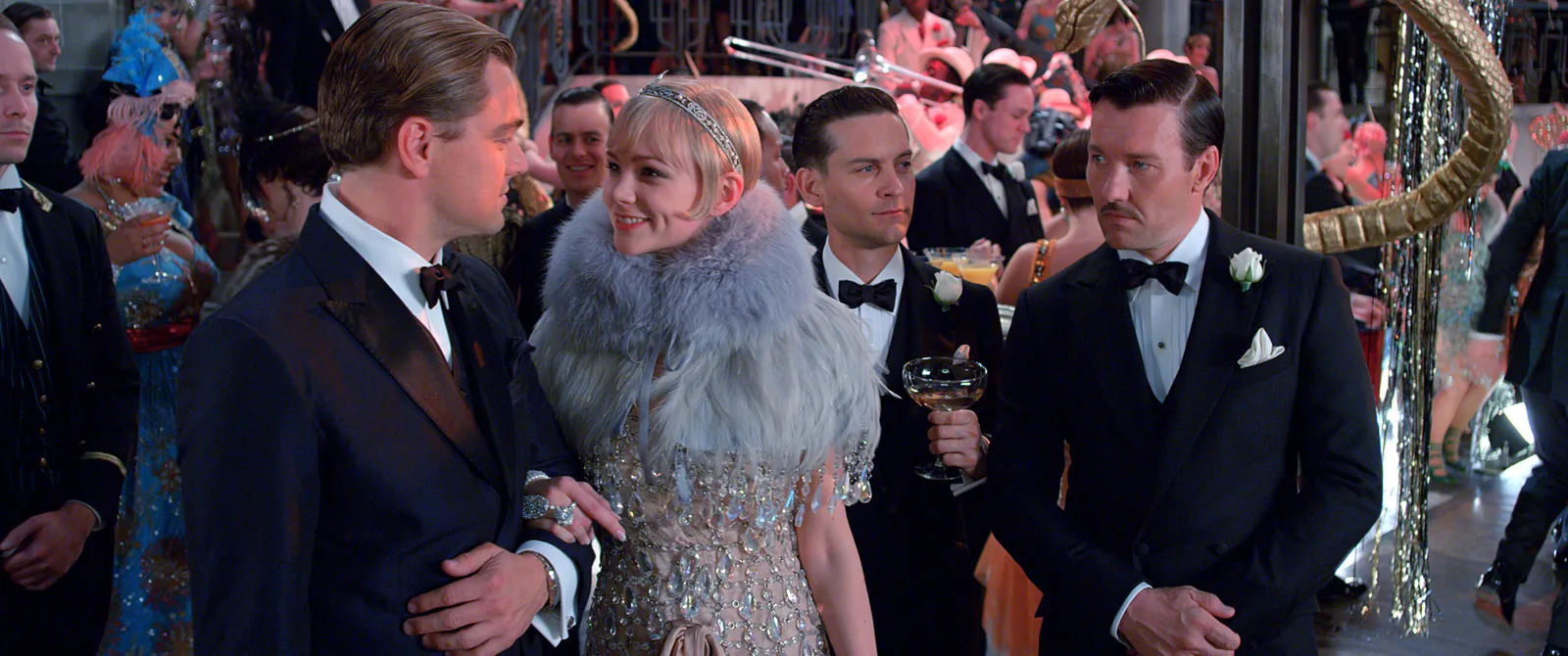 A party scene from The Great Gatsby featuring Daisy, Gatsby, Nick and Tom. Daisy is wearing a blue shawl and them are wearing suits and clutching cocktail glasses.