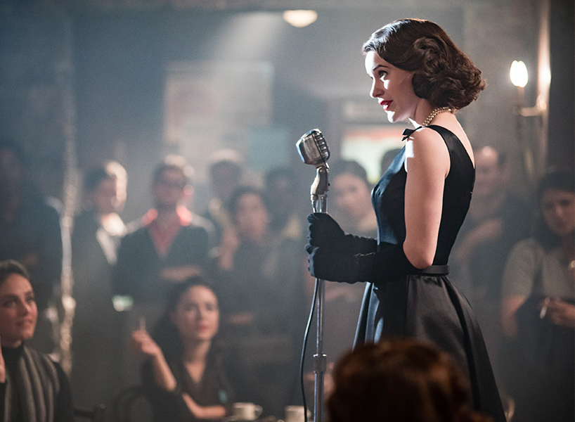Mrs Maisel dressed in a black dress and holding the mic at a event full of people.