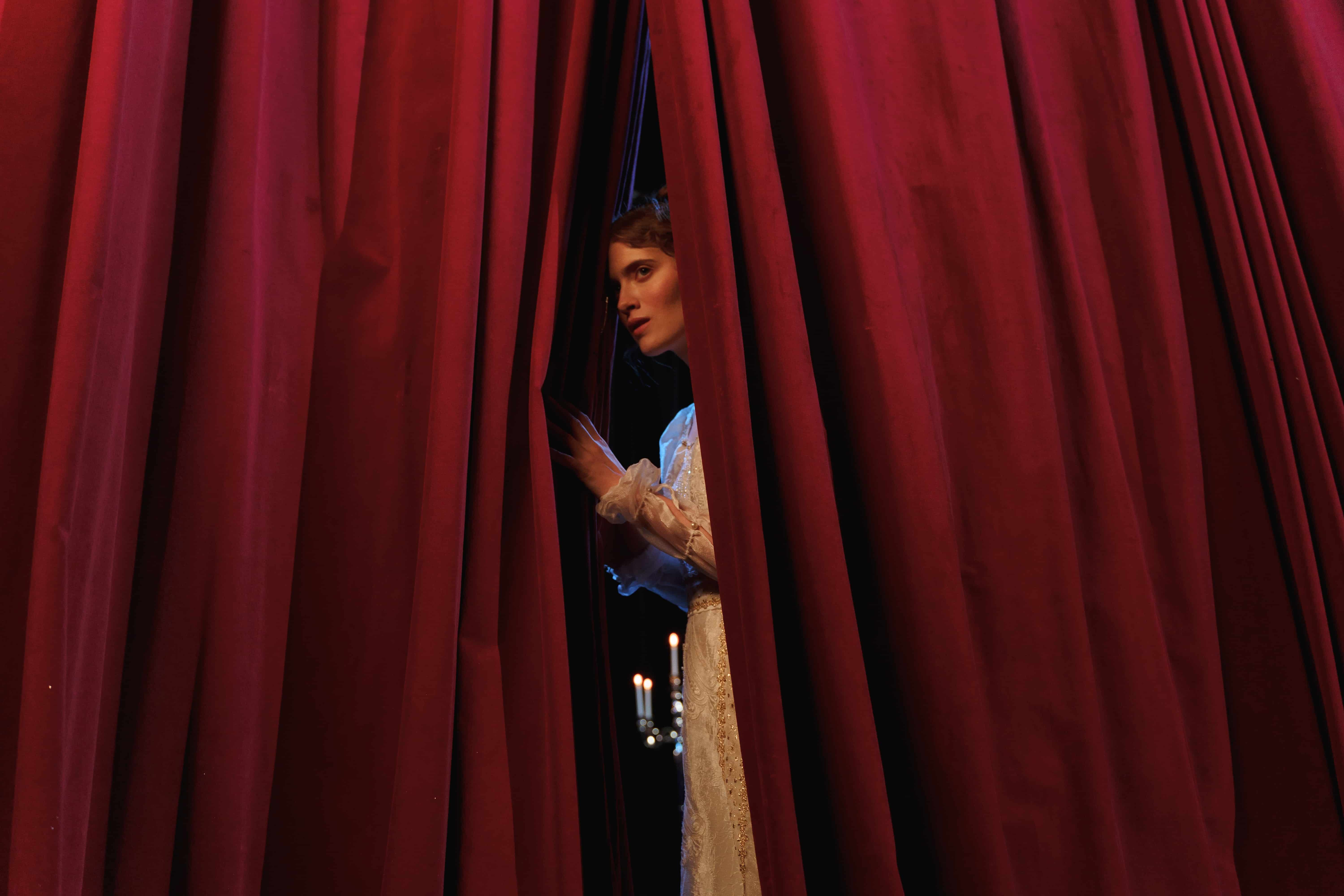 Woman stands behind red curtain.