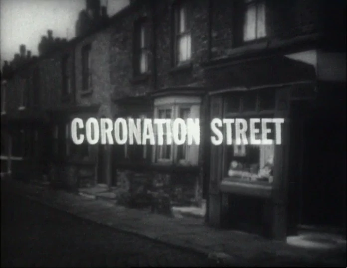 The original black and white title card of Coronation Street, the longest running soap opera in history. It depicts a suburban street in the North of England with pay windows and the all caps words Coronation Street