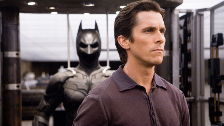 Christian Bale as a pensive Bruce Wayne staring into the distance with his Batman costume behind him